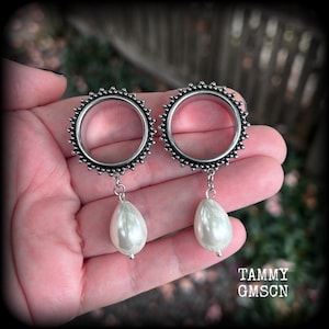 May include: A pair of circular silver-toned ear tunnels featuring a textured, beaded outer edge and a dangling, teardrop-shaped white faux pearl charm suspended from a small metal link at the base of each circular frame.