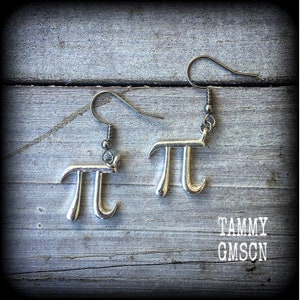 May include: A pair of silver earrings shaped like the mathematical symbol pi. The earrings are hanging from a wooden surface.