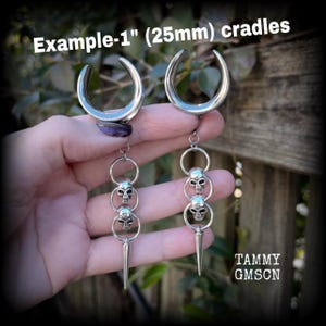 Op de afbeelding: Een paar zilverkleurige, halvemaanvormige oorsieraden van 25 mm. Elk sieraad heeft een hangende ketting met twee kleine schedelbedels en een puntige stekel aan de onderkant. De tekst "Example-1" (25mm) cradles" is in de afbeelding te zien.