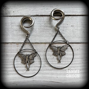 May include: A pair of silver-toned ear gauges with a spiral design. Each gauge is connected to a black chain that suspends a silver moth pendant within a black circular frame. The background is a white wooden surface.