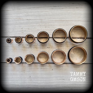May include: A collection of ear gauges in various sizes. The gauges have a gold-colored interior, a black band, and a copper-colored exterior. The gauges are arranged on a weathered, light-colored wooden surface. The text "TAMMY GMSCN" is visible.