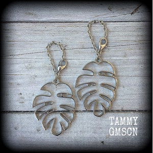 May include: Two silver-coloured Monstera leaf charms with chain and clasp attachments. The charms are displayed on a weathered, grey wooden surface. The text "TAMMY GMSCN" is printed in white.