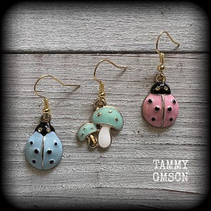 May include: Three pairs of dangle earrings with gold-colored hooks. One pair features a light blue ladybug design with black spots. Another pair has a light blue mushroom design with a white stem. The third pair has a pink ladybug design with black spots. The earrings are on a wooden background.