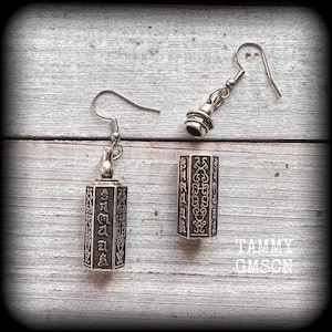May include: A pair of silver earrings with a hexagonal pendant featuring an intricate design. The pendant has a textured surface and appears to be made of metal. The earrings are hanging from a silver hook.