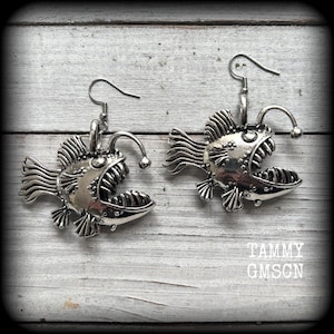 以下が含まれることがあります： 銀色のチョウチンアンコウのイヤリングのペア。各イヤリングは、開いた顎、ひれ、電球のようなルアーを備えた詳細な魚のデザインが特徴です。「TAMMY GMSCN」というテキストが表示されます。