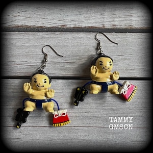 May include: A pair of dangle earrings featuring miniature sumo wrestler figurines with tan skin and dark blue mawashi belts. Each figure is attached to a silver-toned hook and holds a small black fan and a red and white decorative accessory.
