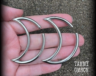 May include: Two polished silver-toned crescent moon-shaped body jewelry pieces with a smooth, reflective finish. Each piece features a curved, open design with pointed ends. The text TAMMY GMSCN is visible in the lower right corner of the image.