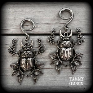 May include: A pair of silver-coloured, ornate earrings with a scarab beetle design. Each earring has a curved top and a detailed beetle pendant with leaf and branch accents. The text "TAMMY GMSON" is visible.