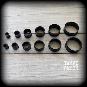 May include: A collection of black ear gauges in various sizes, arranged on a light wood-grain surface. The gauges include both flared and tunnel styles, showcasing a range of diameters. The image is well-lit, highlighting the smooth, dark finish of the jewelry.