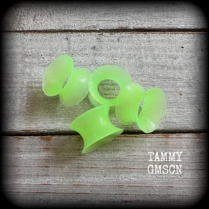 May include: Several neon green ear gauges are arranged on a weathered wooden surface. The gauges have a flared design and a smooth, translucent appearance. The text "TAMMY GMSCN" is visible in the lower right corner.