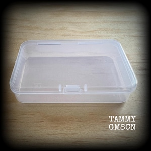 Può includere: Una scatola di plastica trasparente, rettangolare, con coperchio incernierato e chiusura. La scatola è vuota e poggia su una superficie di legno. Il testo "TAMMY GMSCN" è stampato in bianco in basso a destra.