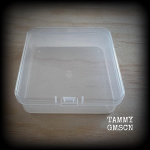 May include: A clear, rectangular plastic storage box with a hinged lid. The box is empty and sits on a wooden surface. The text "TAMMY GMSCN" is printed on the lower right corner.