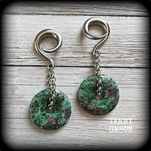 May include: A pair of silver-toned spiral earrings with a chain and a round, green and red stone pendant. The earrings are displayed on a light wood-grain surface. The text "TAMMY GMSCN" is visible in the lower right corner.