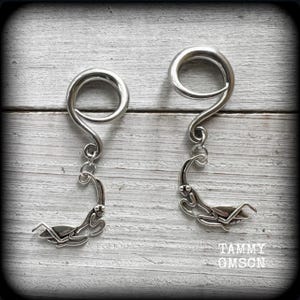 May include: A pair of silver-toned ear gauges with a spiral design and a dangling praying mantis charm. The gauges are displayed on a light-colored, textured surface. The text "TAMMY OMSCN" is visible.