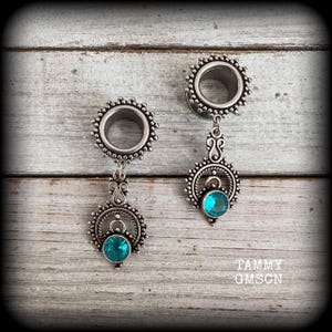 Op de afbeelding: Een paar zilverkleurige oortunnels met een cirkelvormig ontwerp en een kralenrand. Elke tunnel heeft een hangend decoratief element met een ronde, helder turquoise edelsteen in een filigraan zetting. De tekst TAMMY GMSCN is zichtbaar.
