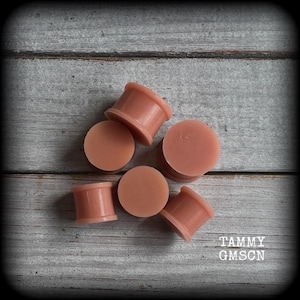 May include: A collection of five smooth, solid, light peach-coloured ear plugs with a cylindrical shape and flared edges. The items are arranged in a cluster, displaying a uniform matte finish across their surfaces. The text TAMMY GMSCN is visible.