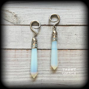 May include: Two silver earrings with a spiral design and long, pale blue, crystal-like pendants. The pendants are pointed at the base. The earrings are presented on a light wood surface. The text "TAMMY GMSGN" is visible.