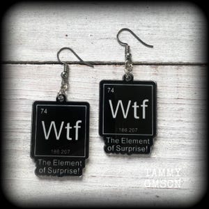 May include: A pair of black earrings with a white border, each displaying the word "Wtf" in white, alongside the numbers "74" and "186 207". The text "The Element of Surprise!" is below the main design. The earrings hang from silver-coloured hooks.