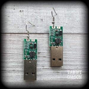 May include: A pair of earrings made from repurposed circuit boards with USB connectors. The circuit boards are green with black components and silver connectors. The earrings are attached to silver hooks.