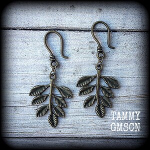 May include: A pair of bronze-toned metal earrings featuring a detailed leaf branch design. Each earring hangs from a curved hook and displays a central stem with multiple small, textured leaves extending outward. The text TAMMY GMSCN is visible.