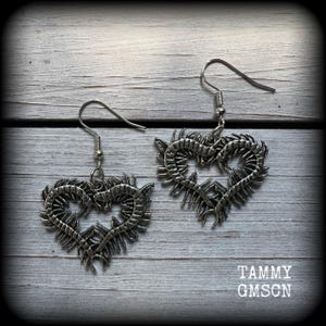 May include: A pair of silver-toned heart-shaped earrings. Each earring features a detailed, spiky design around the heart's outline, with a fish hook style ear wire. The earrings are displayed on a light gray wooden surface.