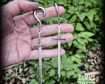 May include: A pair of silver-toned ear weights featuring a curved hook design at the top, each suspending a detailed metal sword pendant. The swords have a crossguard, a textured grip, and a long, straight blade, hanging vertically from the hooks.