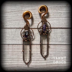 Può includere: Due dilatatori per orecchie a spirale color oro con pendenti decorativi. Ogni pendente presenta un cristallo trasparente, una gemma viola e una cornice in metallo color rame. I dilatatori sono esposti su una superficie di legno invecchiato.