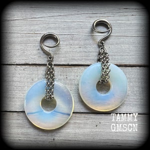 May include: A pair of opalite stone earrings with a silver-tone chain and hook design. The earrings feature a circular opalite stone with a hole in the center, suspended from a silver-tone hook. The earrings are displayed on a light wood surface.