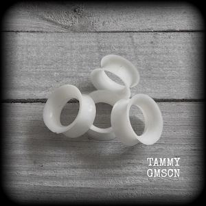May include: Several white, circular ear gauges are arranged on a gray wooden surface. The gauges have a smooth, rounded design. The text "TAMMY GMSON" is printed in white at the bottom right corner.