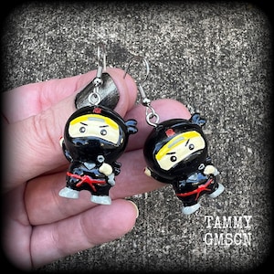 May include: A pair of black and yellow ninja earrings with red accents. The earrings are made of polymer clay and have silver hooks.