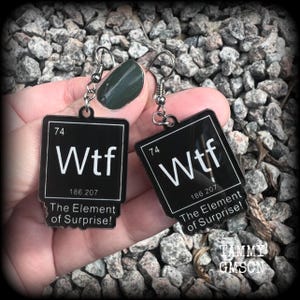 May include: Two black earrings with the text "Wtf" in white, styled like a periodic table element. The earrings also feature the text "The Element of Surprise!" and are suspended from silver hooks.