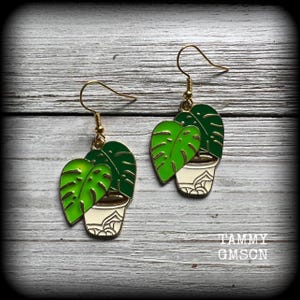 Op de afbeelding: Goudkleurige oorbellen met een groen monstera blad ontwerp en een witte en gouden plantenpot. De oorbellen hebben een haaksluiting. De tekst "TAMMY GMSCN" is rechtsonder zichtbaar.