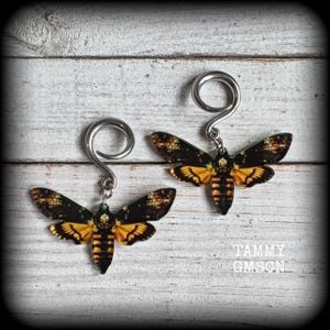 May include: A pair of silver-toned spiral earrings with Death's Head moth pendants. The moth pendants feature black and yellow wings with intricate patterns. The earrings are displayed on a white wooden surface.