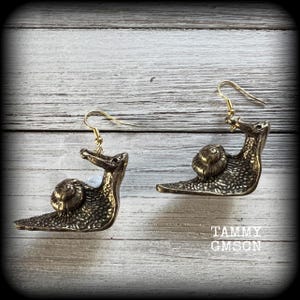 May include: A pair of bronze-colored snail earrings with gold-colored ear wires. The earrings are detailed with textured shells and bodies. The text "TAMMY GMSON" is visible in the lower right corner. The earrings are displayed on a light wood-grain surface.