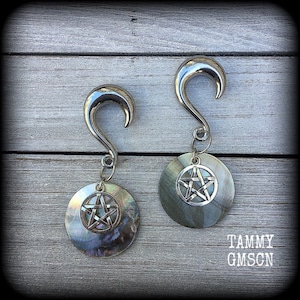 May include: A pair of silver-toned ear gauges with a curved hook design. Each gauge features a circular pendant with a pentagram symbol. The pendants have a pearlescent, iridescent finish. The text "TAMMY GMSON" is visible in the bottom right corner.
