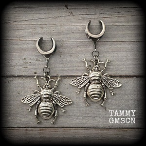 Peut inclure: Une paire d'écarteurs d'oreille argentés avec des breloques abeilles pendantes. Les breloques abeilles ont des ailes et des segments de corps détaillés. Les jauges sont suspendues à un poteau incurvé en forme de fer à cheval. Fond en bois gris vieilli.