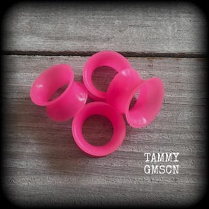 May include: Four bright pink, open-ended ear gauges are arranged on a weathered, gray wooden surface. The gauges are made of a smooth material and have a flared design. The text "TAMMY GMSCN" is in the lower right corner.