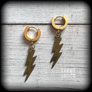 May include: A pair of gold-colored hoop earrings with lightning bolt charms. The earrings have a hinged closure and a textured, antique brass finish on the lightning bolt pendants. The earrings are displayed on a white wooden surface.