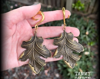 May include: A pair of brass-toned ear weights shaped like oak leaves with a textured, pitted surface. Each leaf hangs from a smooth, curved hook designed for ear stretching. The text TAMMY GMSCN is visible in the bottom right corner.