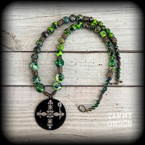May include: A handcrafted necklace featuring a black pendant with a white design, paired with green and black speckled beads. The necklace includes bronze-colored metal accents and a toggle clasp. The artist's name, Tammy Gmson, is visible in the image.