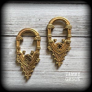 May include: A pair of gold-coloured earrings with an elaborate gothic arch design. Each earring has a decorative top, a central quatrefoil, and a pointed base. They are displayed on a light wood-grain surface. The text "TAMMY GMSON" is visible.