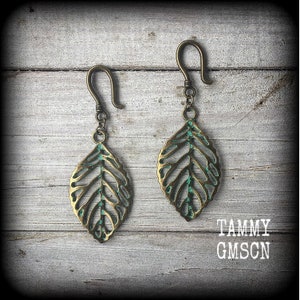 May include: A pair of bronze-coloured leaf earrings with a verdigris patina. The earrings feature a leaf vein design and a hook closure. They are displayed on a weathered wooden surface. The text "TAMMY GMSCN" is visible.
