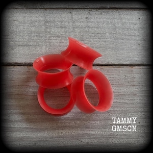 May include: Several red silicone ear gauges are arranged on a weathered wooden surface. The gauges are circular with flared edges. The text "TAMMY GMSCN" is visible in the lower right corner. These are body jewelry.
