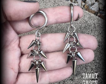Puede incluir: Un par de pendientes plateados con un diseño único. Cada pendiente presenta una serie de púas en forma de cono suspendidas de un gancho curvo. Los pendientes se sostienen en una mano, mostrando sus intrincados detalles y su estética atrevida. El texto "TAMMY GMSCN" es visible.