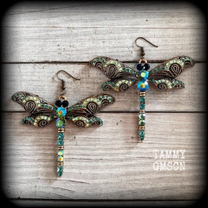 May include: A pair of dragonfly earrings featuring antique bronze-toned metal filigree wings encrusted with small green and iridescent crystals. The bodies are composed of a vertical row of round green crystals. The text TAMMY GMSCN is visible.