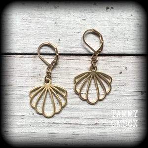 May include: A pair of gold-toned metal earrings featuring a cutout seashell design. Each earring is suspended from a hinged leverback closure. The shell charms have a symmetrical, fan-like shape with open spaces between the metal ribs.