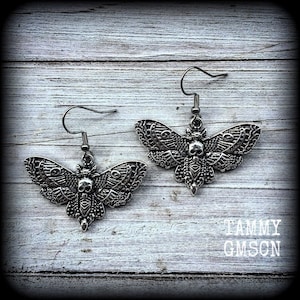 May include: A pair of silver-toned moth earrings. Each earring features a detailed moth design with a skull at the center. The moth's wings are intricately patterned. The earrings hang from a simple hook.