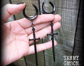 May include: A pair of black ear gauges featuring a crescent moon shape with a dangling sword pendant attached by a small metal ring. The jewellery has a smooth, polished finish and includes the text TAMMY GMSCN in the bottom right corner.