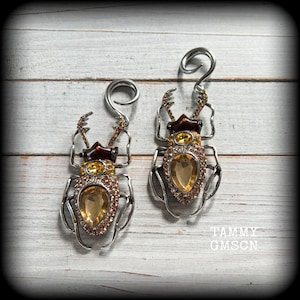 May include: A pair of silver-toned beetle-shaped ear gauges with spiral tops. Each insect features a dark brown head, a central amber-coloured teardrop crystal, and a body encrusted with small orange and clear rhinestones. Text reads TAMMY GMSCN.