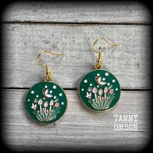May include: A pair of gold-colored earrings with a circular, dark green pendant. Each pendant features a design of mushrooms, butterflies, and white dots. The earrings have a gold-colored hook.